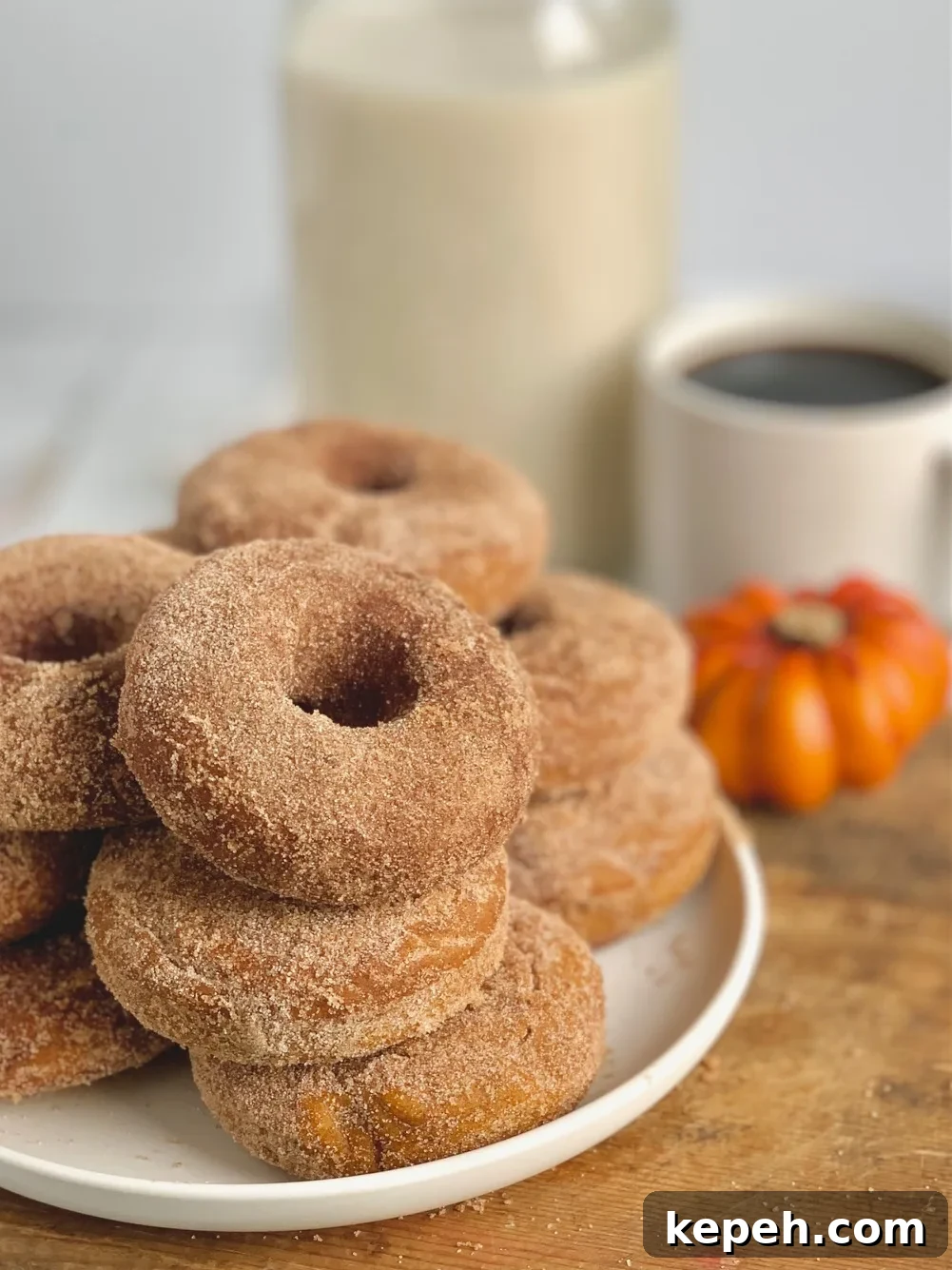 Several donuts stacked on top of each other on a white plate next to a cup of coffee and a decorative pumpkin.