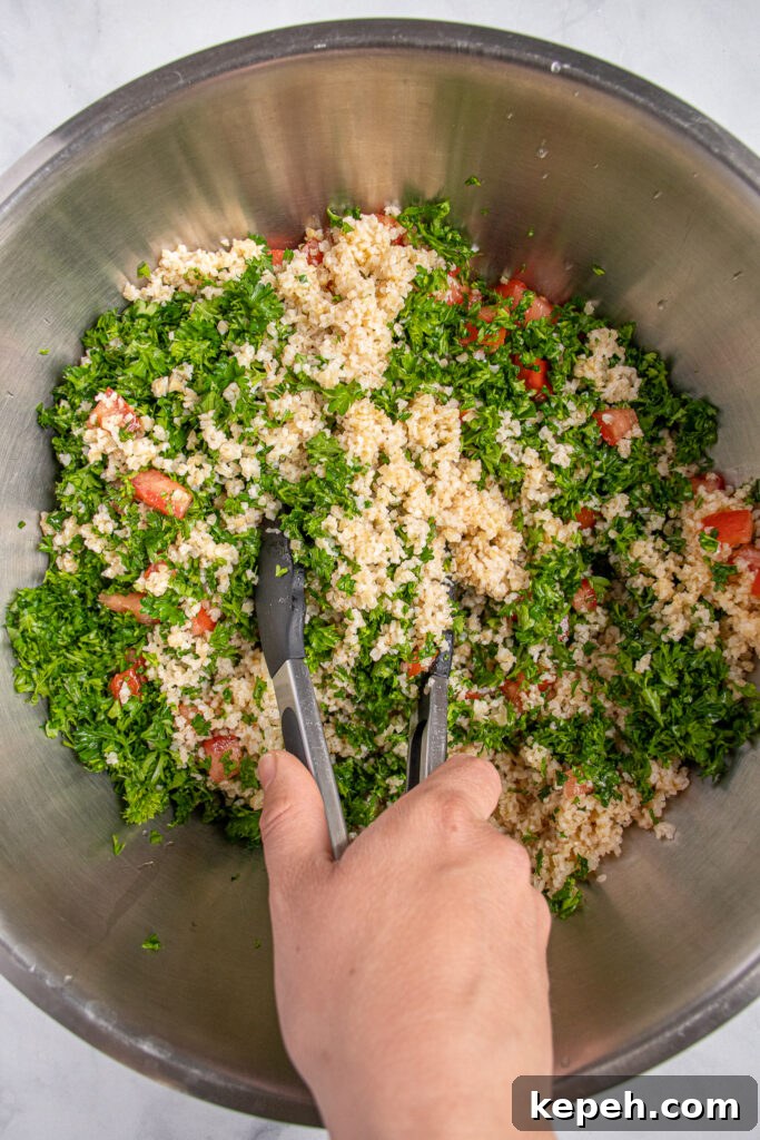 A person using tongs to mix the ingredients of tabbouleh salad together including parsley, bulgur, and tomatoes.
