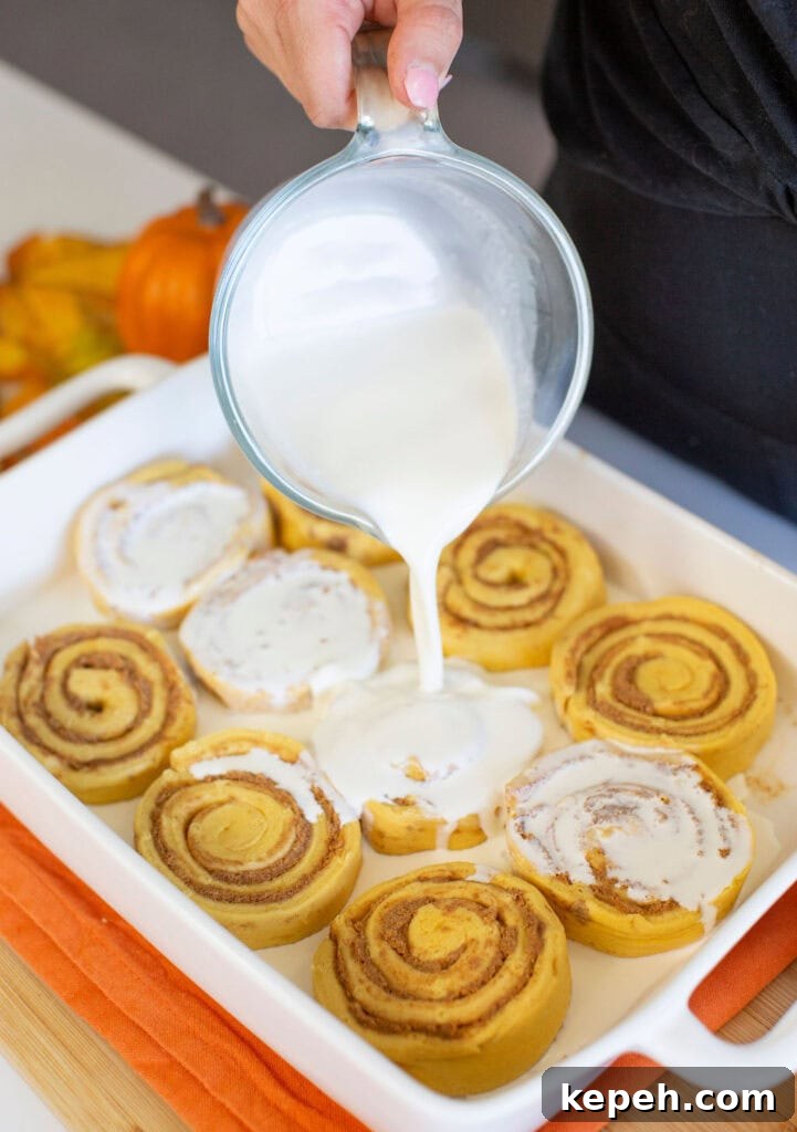 Pumpkin Cinnamon Rolls Made with Rich Whipping Cream 5 A person pours creamy icing over freshly baked cinnamon rolls in a white rectangular dish. The dish is set on an orange cloth. A small pumpkin is visible in the background, suggesting an autumn setting.