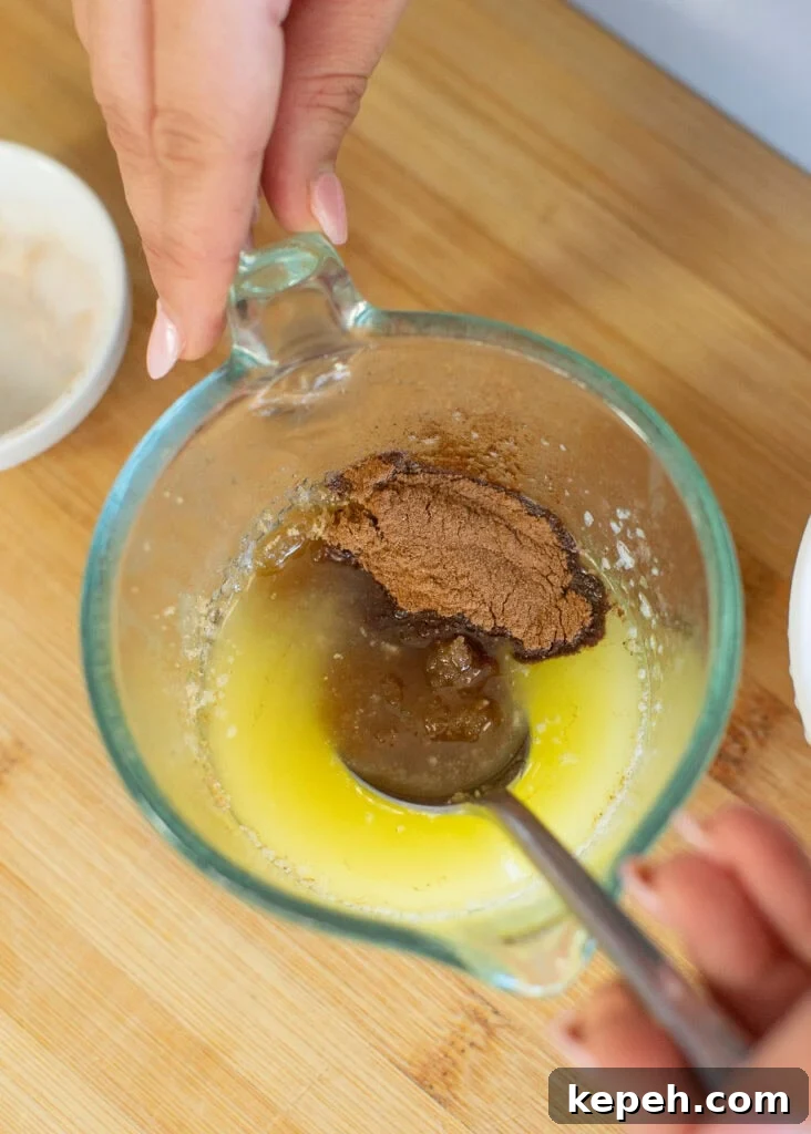 Pumpkin Cinnamon Rolls Made with Rich Whipping Cream 6 Close-up of hands holding a glass measuring cup with melted butter and a spoon mixing in brown sugar and cinnamon. The cup is on a wooden surface with a small bowl nearby.