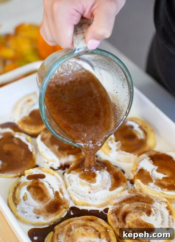 Pumpkin Cinnamon Rolls Made with Rich Whipping Cream 7 A person pours a cinnamon sugar mixture over uncooked cinnamon rolls in a white baking dish. The rolls have visible swirls and are topped with drizzles of white icing.