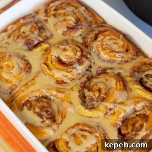 Pumpkin Cinnamon Rolls Made with Rich Whipping Cream 10 A close-up of freshly baked cinnamon rolls in a rectangular white dish, topped with a creamy glaze. A hand holds one side of the dish. The rolls are tightly packed and glistening with the glaze.