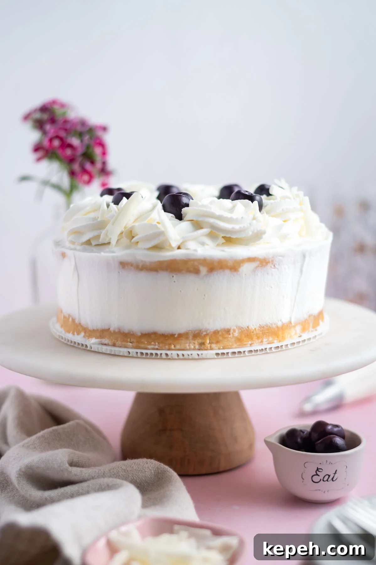 Light and Fluffy White Forest Cake Recipe 18 white forest cake on a cake stand with flowers in the background and a bowl of cherries