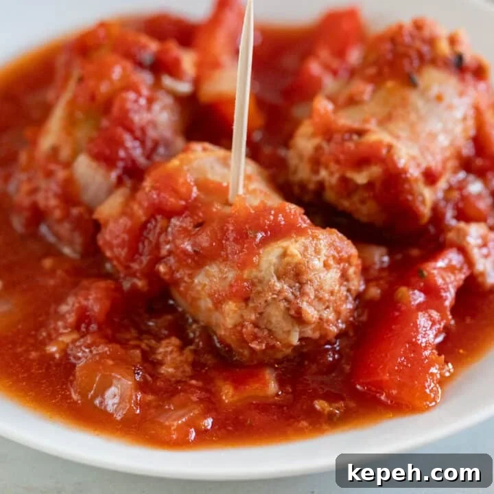 A close up image of a chunk of Italian sausage in red sauce on a white plate with a toothpick sticking out of it.