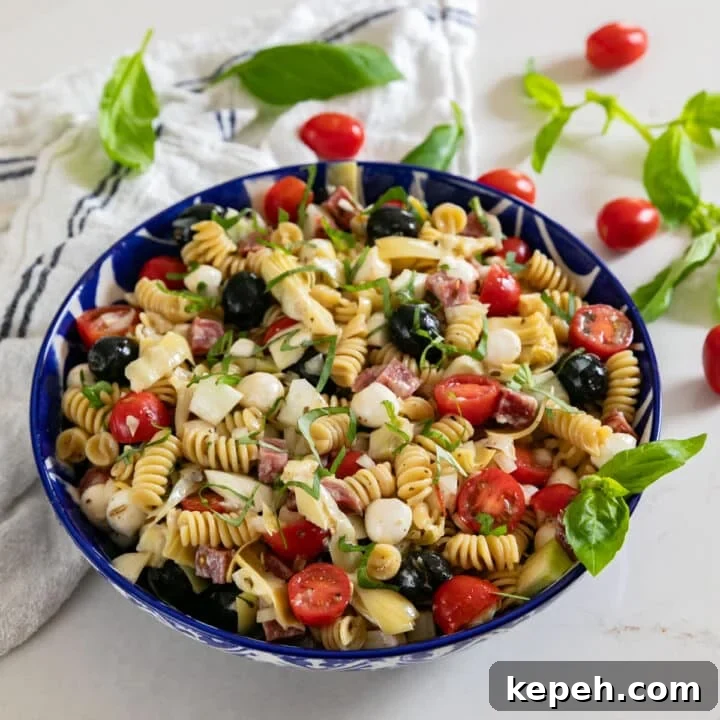 A bowl of pasta salad with a white towel behind it and tomatoes and basil around it.