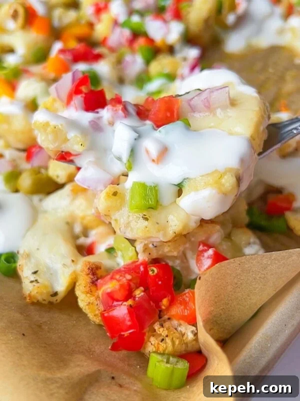 Loaded Cauliflower Nachos with Cheese, Salsa & Avocado 3 A close-up of a fork lifting loaded cauliflower nachos topped with diced red tomatoes, green onions, red onions, melted cheese, and a drizzle of white sauce on a parchment-lined tray.