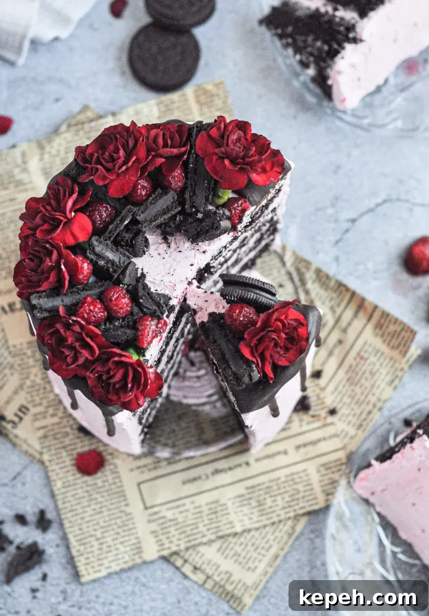 A pink frosted cake with chocolate layers and topped with dark chocolate oreos, raspberries, and bright red flowers. There are two slices taken out of it.