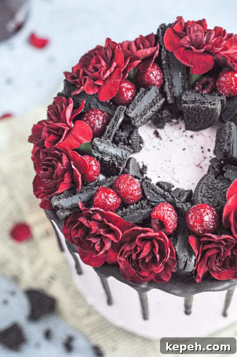 A pink frosted cake with chocolate layers and topped with dark chocolate oreos, raspberries, and bright red flowers.