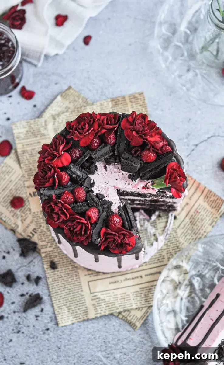 A pink frosted cake with chocolate layers and topped with dark chocolate oreos, raspberries, and bright red flowers.