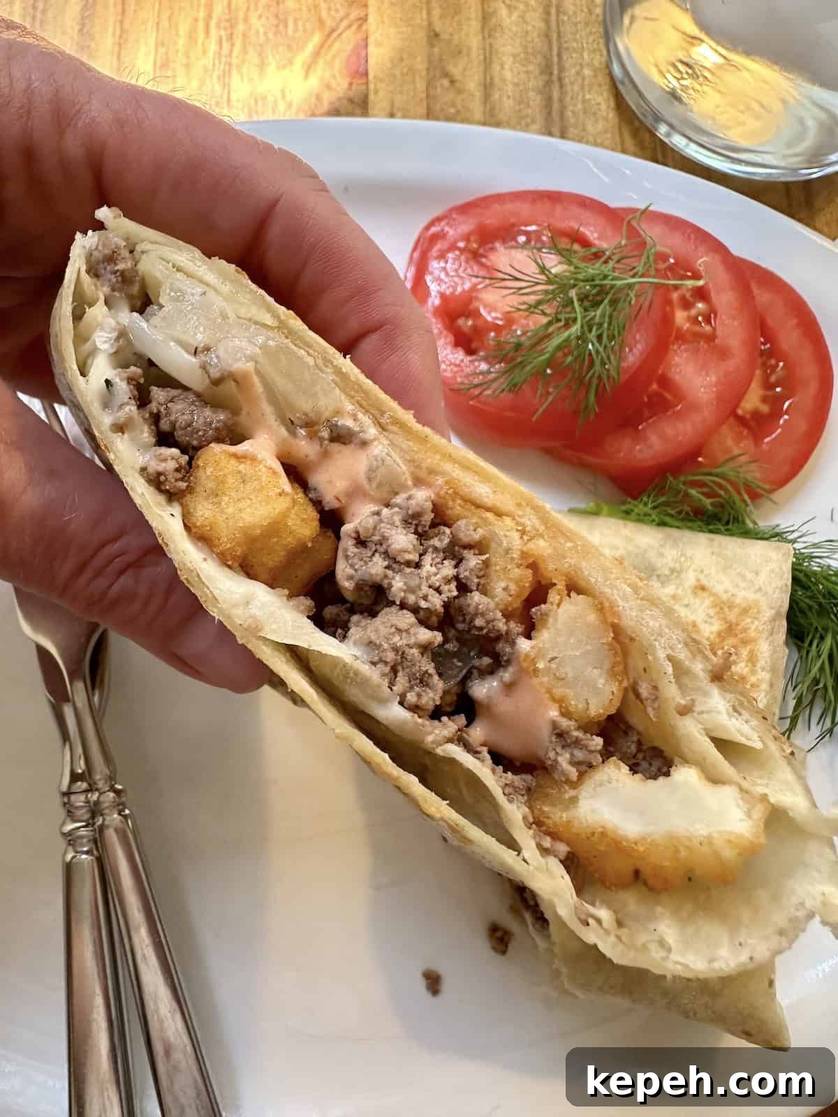 How to Make Authentic French Tacos at Home 2 One half of a French taco held by a hand over a plate with silverware, sliced tomato and dill.