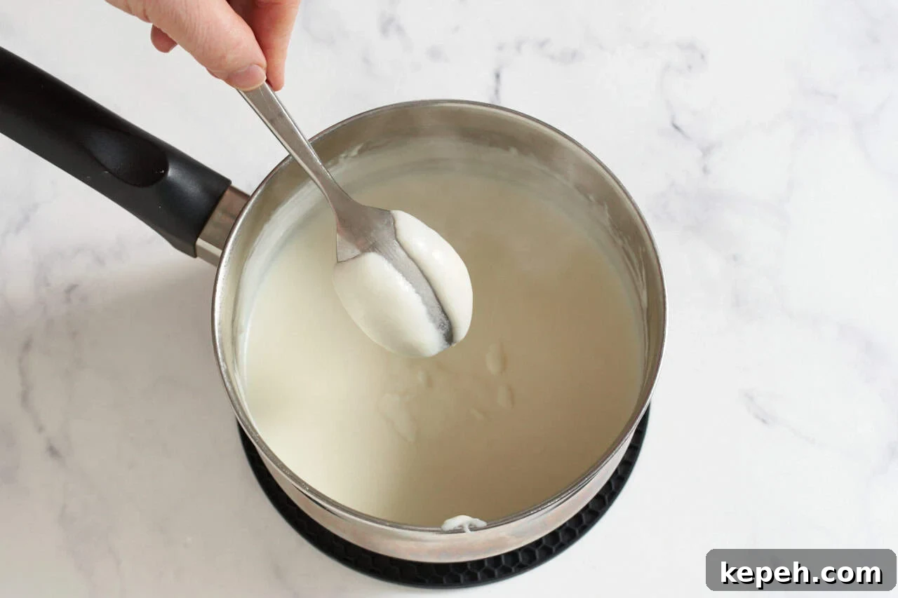 A woman's hand holding a spoon coated with bechamel sauce over a sauce pan of bechamel.