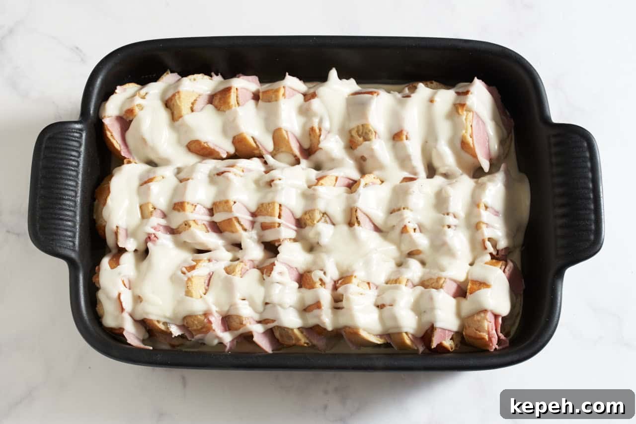 Stacked baguette slices in a black casserole dish layered with ham slices and topped with bechamel sauce.