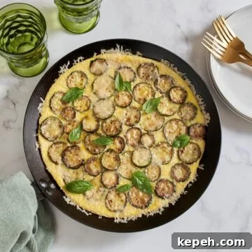 A zucchini frittata in a cast iron pan, two green glasses and a stack of white plates with gold forks on it are above it.