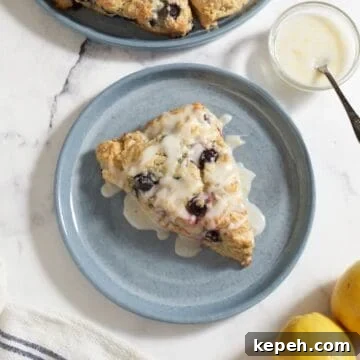 A glazed lemon blueberry scone on a blue plate, a small bowl of lemon glaze is in the upper right corner.