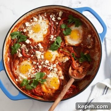 Shakshuka with feta and cilantro in a blue pan with a wooden spoon in it. A blue and white striped towel is on the right.