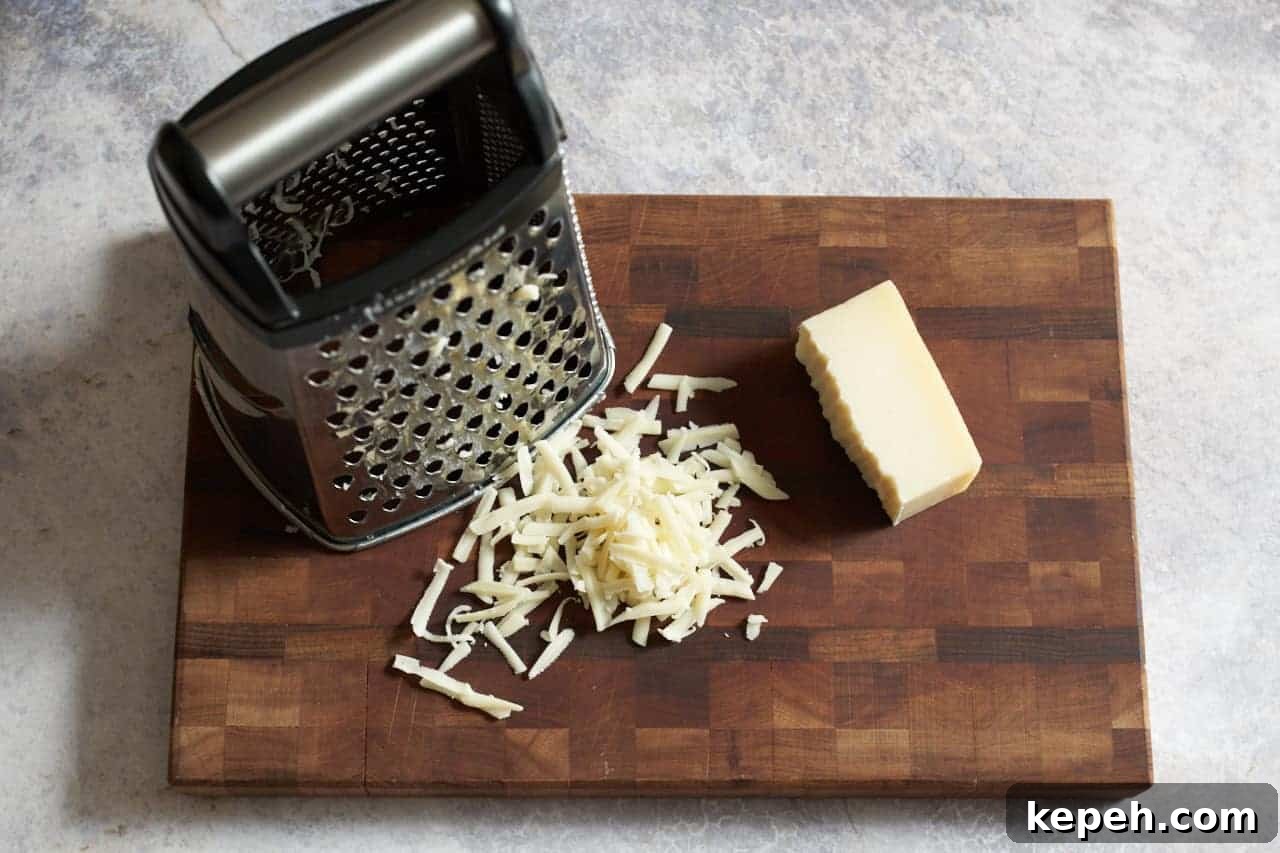 A box grater on a cutting board with grated cheese and a small block of gruyere cheese.