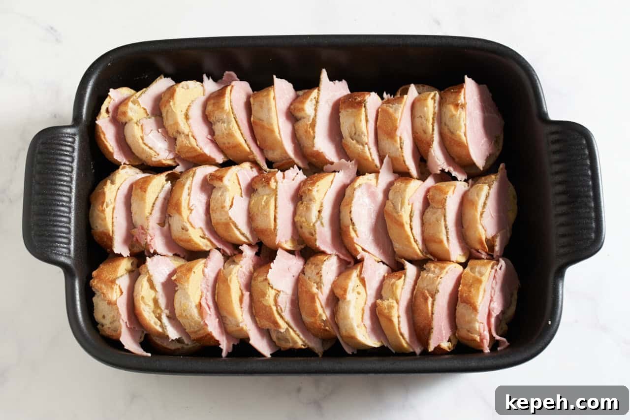 Stacked baguette slices in a black casserole dish layered with ham slices.
