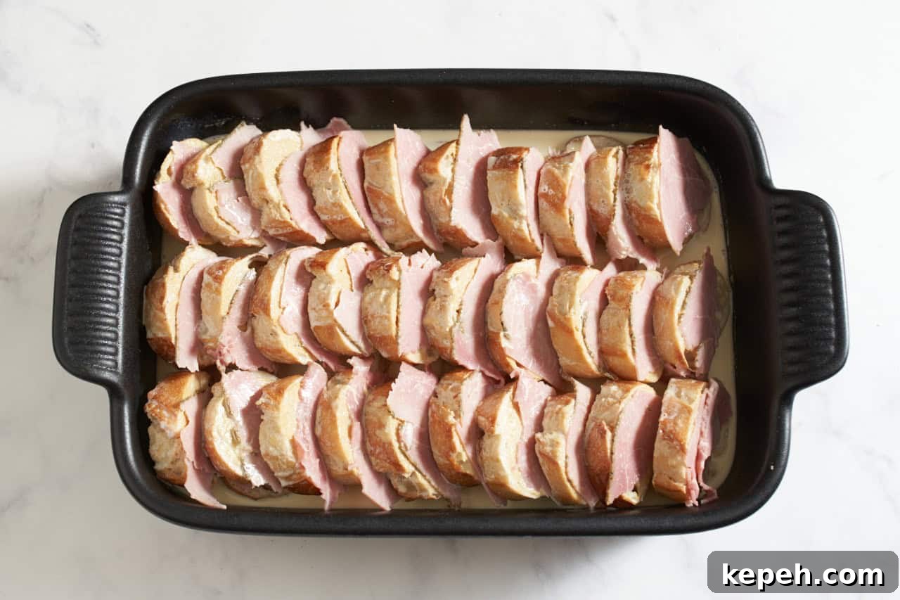 Stacked baguette slices in a black casserole dish layered with ham slices, an egg custard mixture has been poured into the dish.