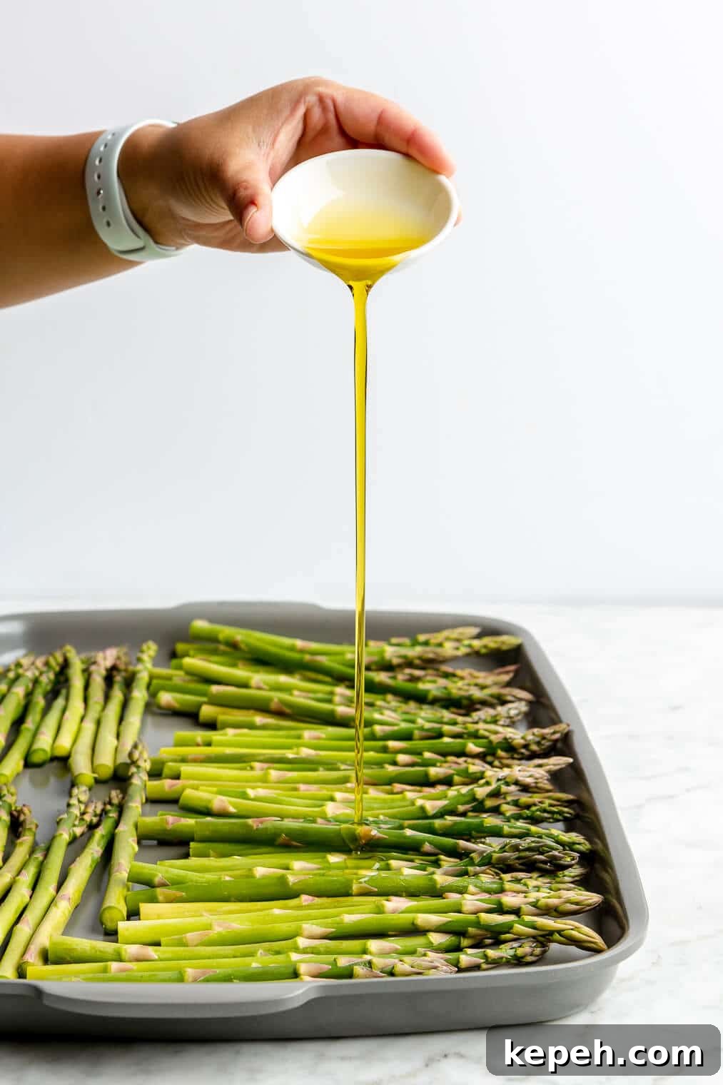 Roasted Asparagus with Lemon Cardamom Cream Sauce 5 A person pouring a small bowl of olive oil over asparagus on a sheet pan.