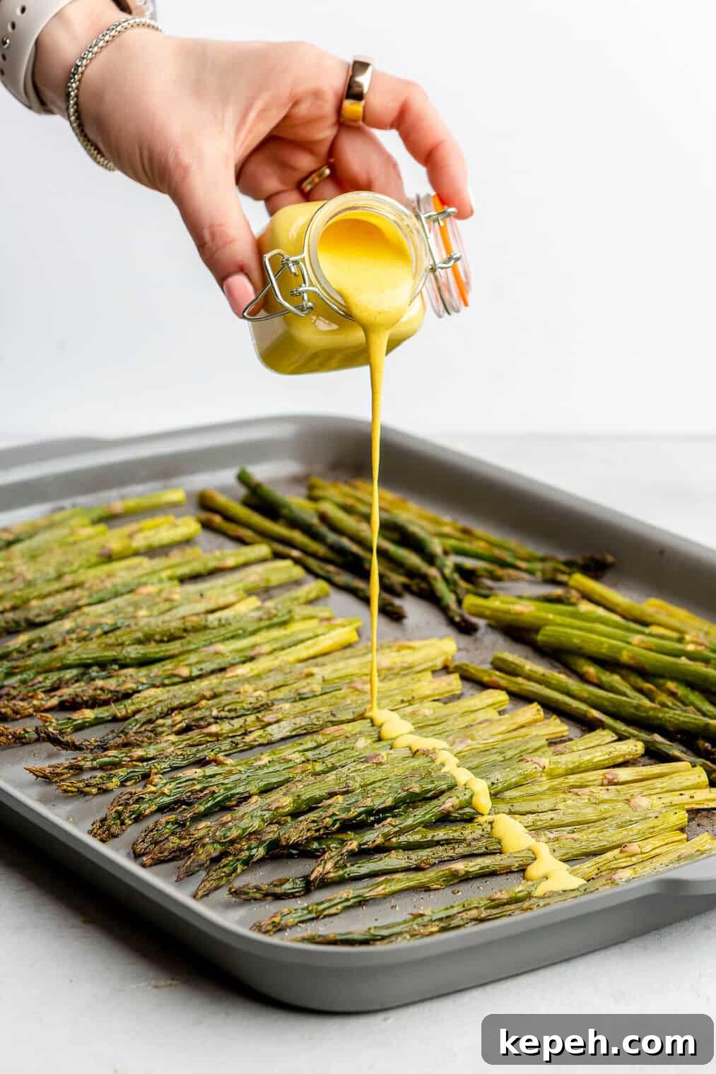 Roasted Asparagus with Lemon Cardamom Cream Sauce 9 A person pouring bright yellow lemon cardamom sauce over a sheet pan of roasted asparagus.