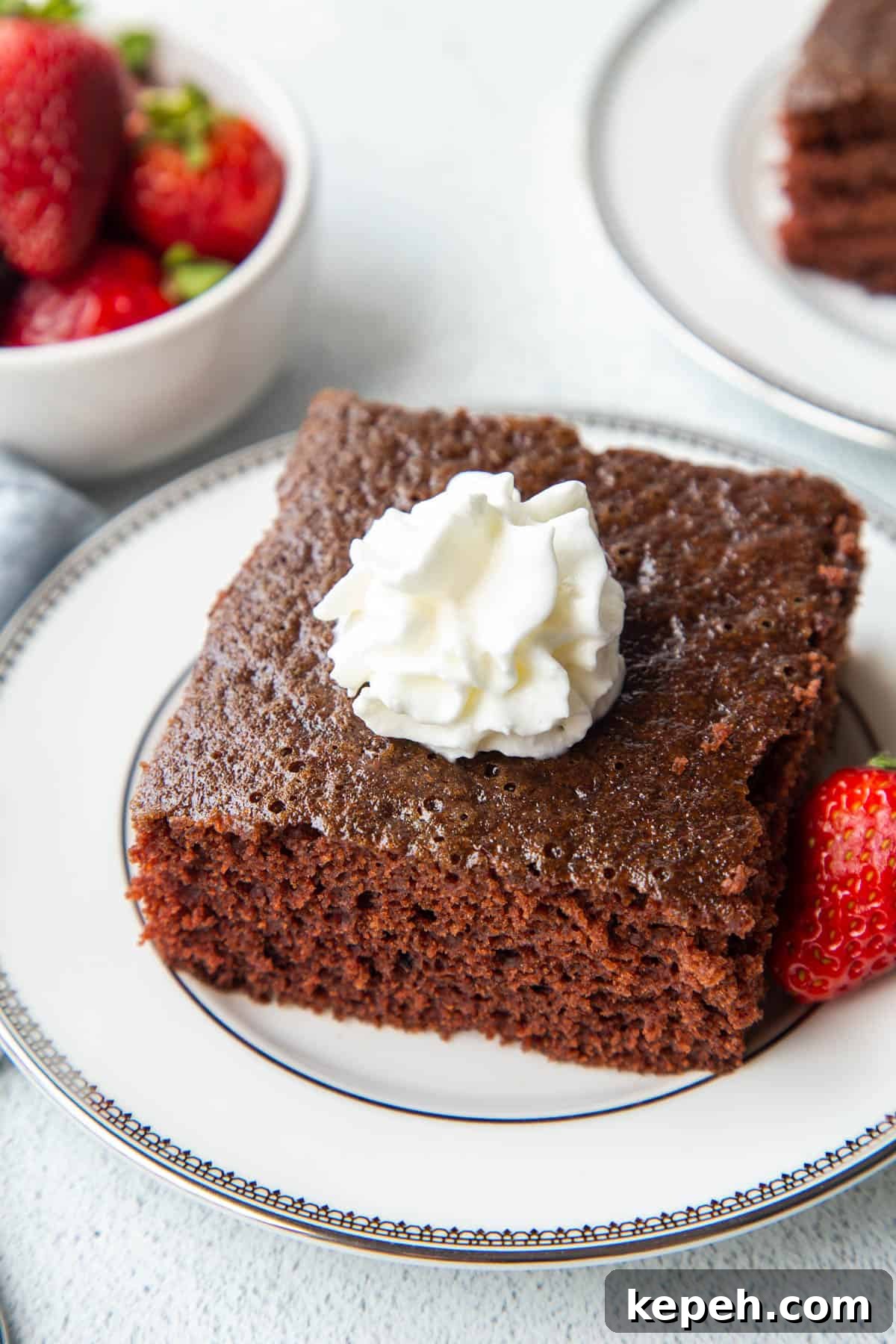 Eggless Wacky Cake Recipe: Moist Chocolate Cake Without Eggs or Milk 2 slice of wacky cake on a white plate, surrounded by strawberries and topped with whipped cream.