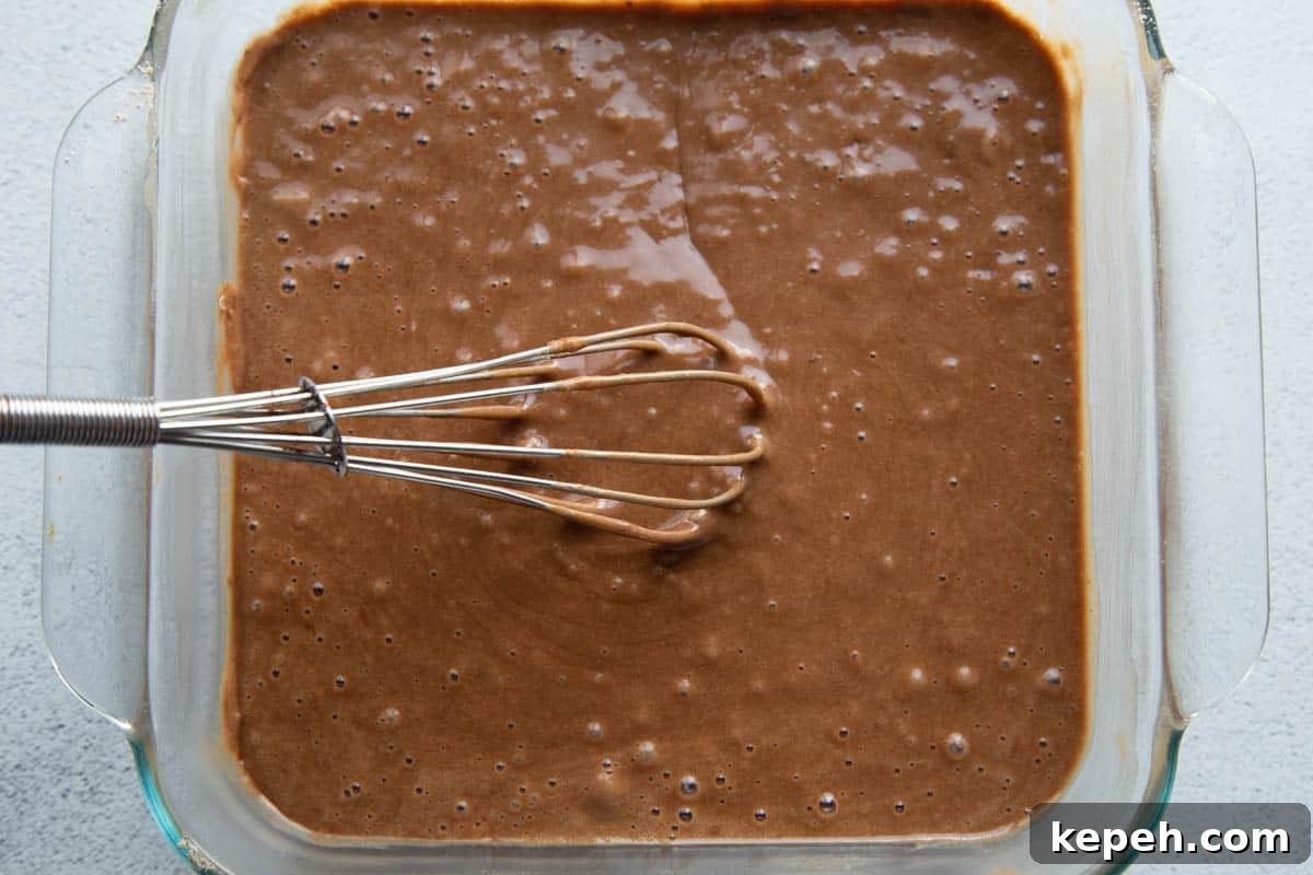 Eggless Wacky Cake Recipe: Moist Chocolate Cake Without Eggs or Milk 5 chocolate cake batter in a square glass disk with a whisk.