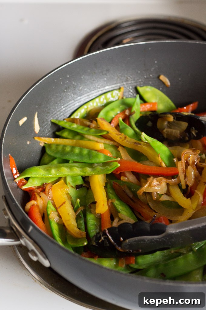 cooked onion and bell pepper slices in a large frying pan with snow peas and a pair of tongs