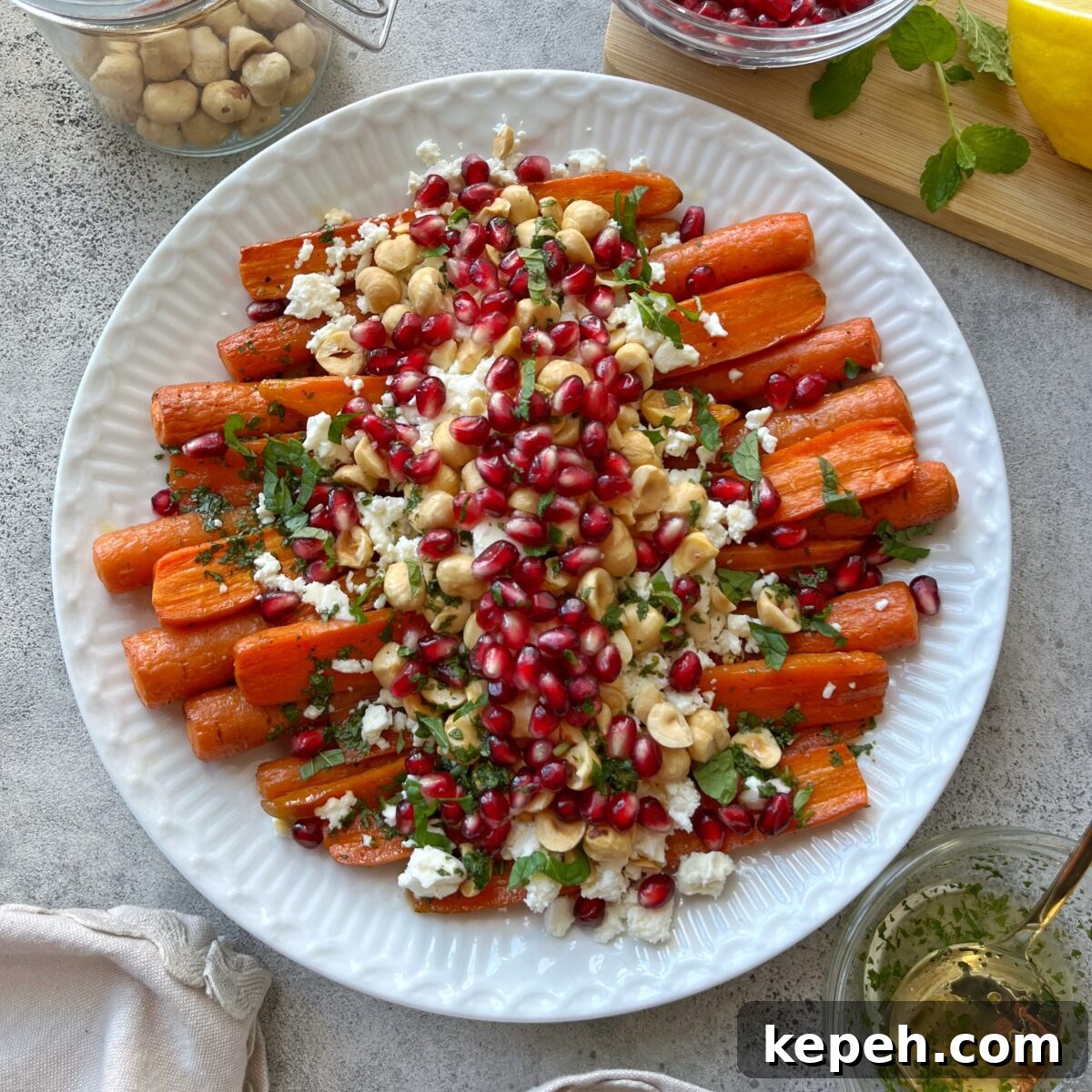 A white plate of Roasted Maple Carrots topped with pomegranate seeds, chopped nuts, crumbled cheese, and fresh herbs, with small bowls of ingredients around it.
