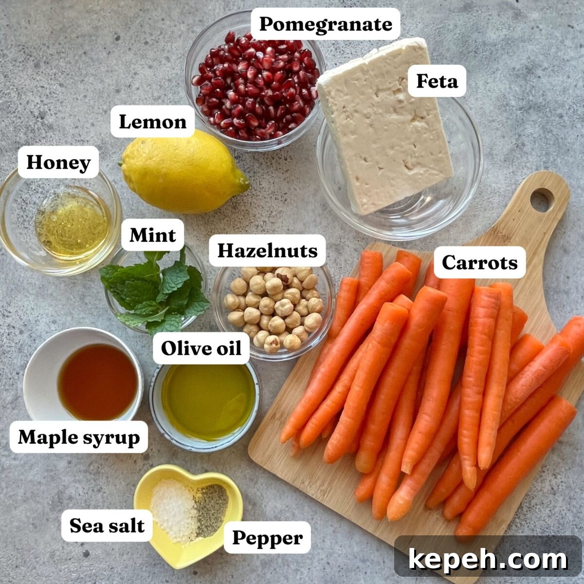 Ingredients for Roasted Maple Carrots are arranged on a surface, including carrots, feta, pomegranate seeds, lemon, honey, mint, hazelnuts, olive oil, maple syrup, sea salt, and pepper.