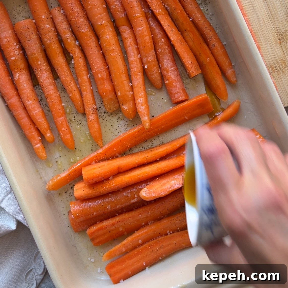 A hand pours maple syrup over raw carrots arranged in a baking dish, sprinkled with salt and pepper.