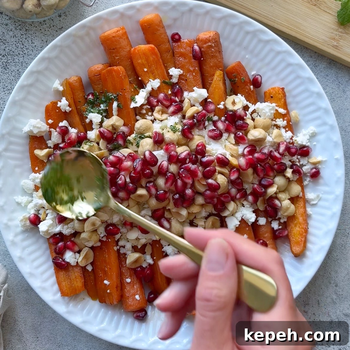 A hand spoons dressing over a plate of Roasted Maple Carrots, topped with feta cheese, hazelnuts, and pomegranate seeds.