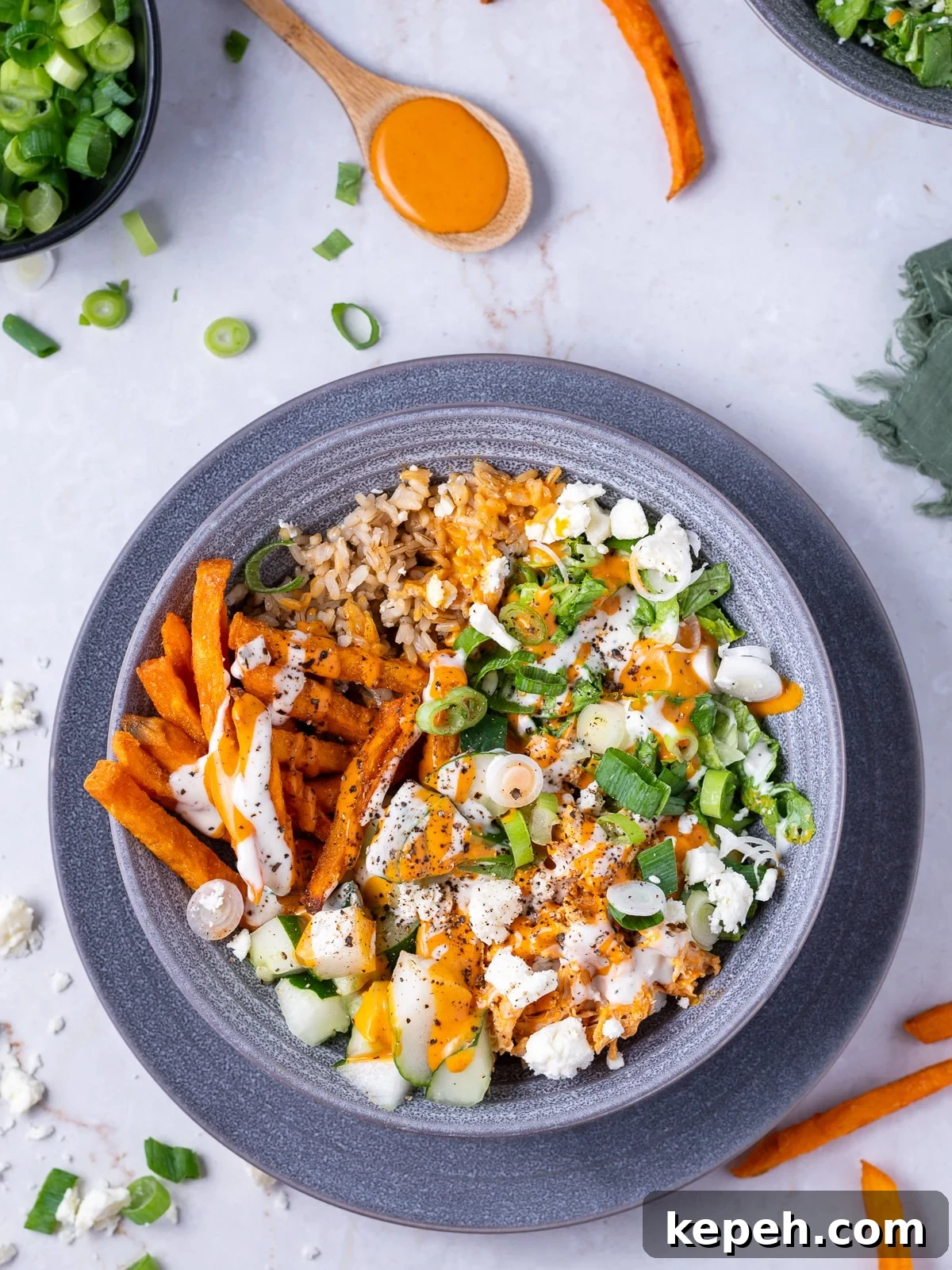 gray bowl with brown rice, sweet potato fries, shredded buffalo chicken, green onions, cucumber, feta, buffalo sauce and ranch dressing