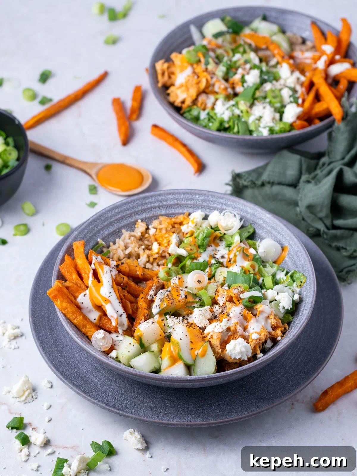 two bowls with brown rice, sweet potato fries, shredded buffalo chicken, green onions, cucumber, feta, buffalo sauce and ranch dressing