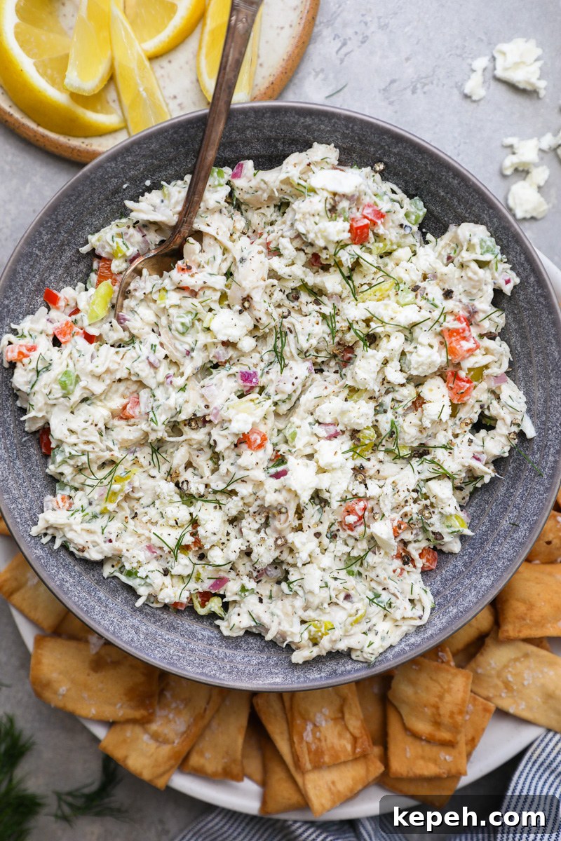 A blue serving bowl filled with feta chicken salad sitting next to pita chips and fresh lemon wedges