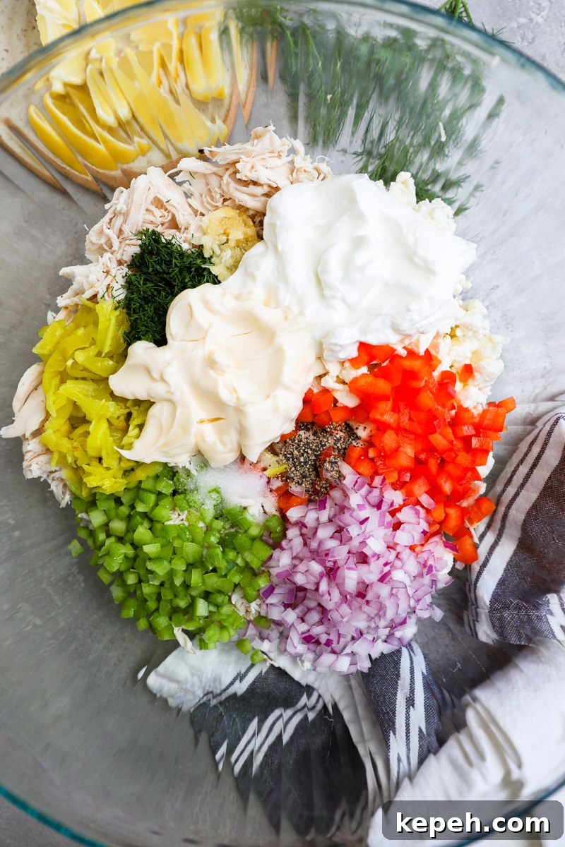 A large mixing bowl filled with shredded chicken, mayonnaise, Greek yogurt, bell pepper, red onion, celery, pepperoncini, fresh dill and garlic