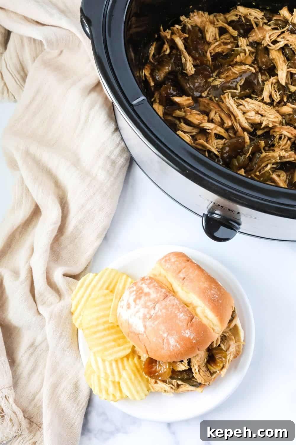 overhead view of a dill pickle chicken sandwich on a plate with potato chips, next to a Crock Pot filled with more of the shredded chicken and pickles.