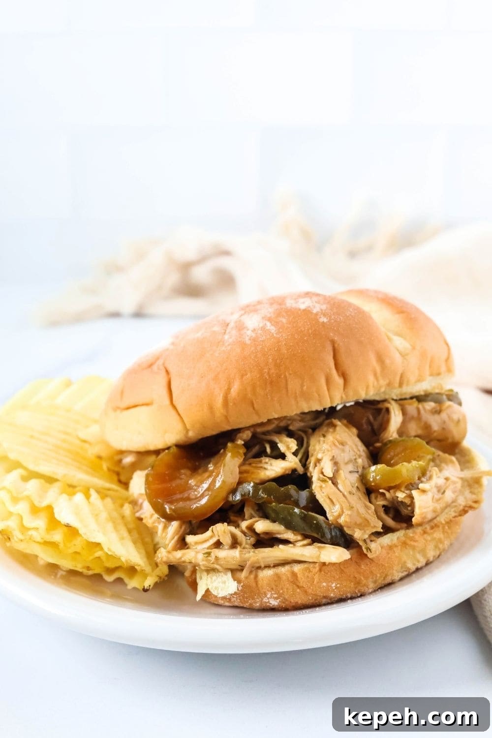 A bun filled with dill pickle chicken for sandwiches, served on a white plate with some potato chips.
