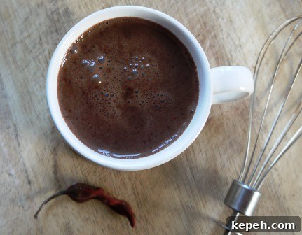 Hot Chilli Chocolate in a white mug beside a chilli and a whisk on a wooden table