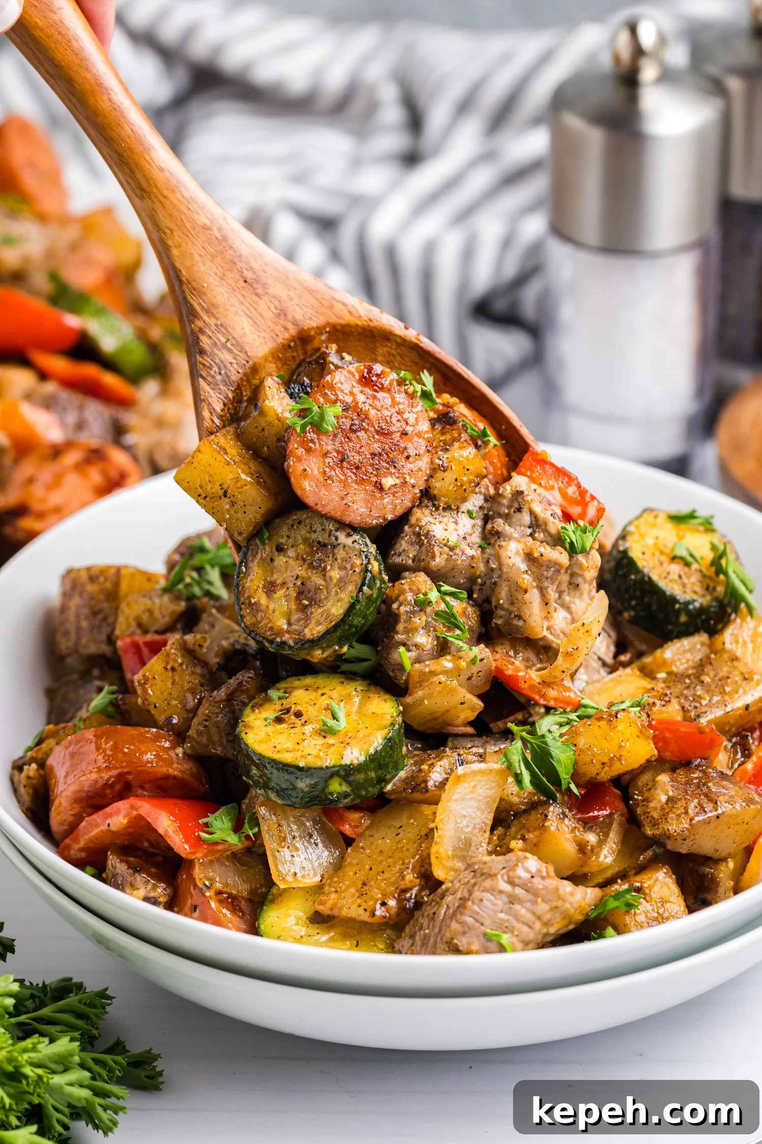 Wooden spoon lifting a serving of sausage, beef, potatoes, zucchini, and peppers from a bowl, garnished with chopped parsley.
