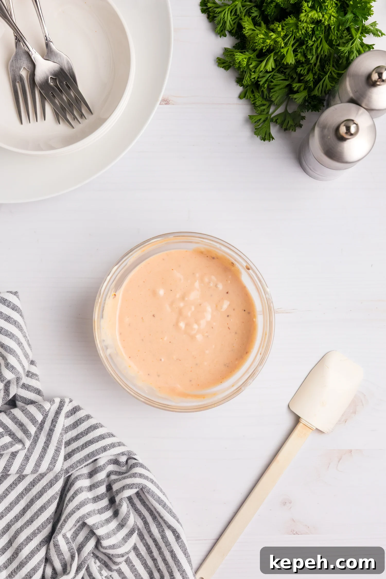 Small bowl of creamy pink sauce on a white surface with a striped towel, spatula, plates and forks, fresh parsley, and salt and pepper shakers nearby.