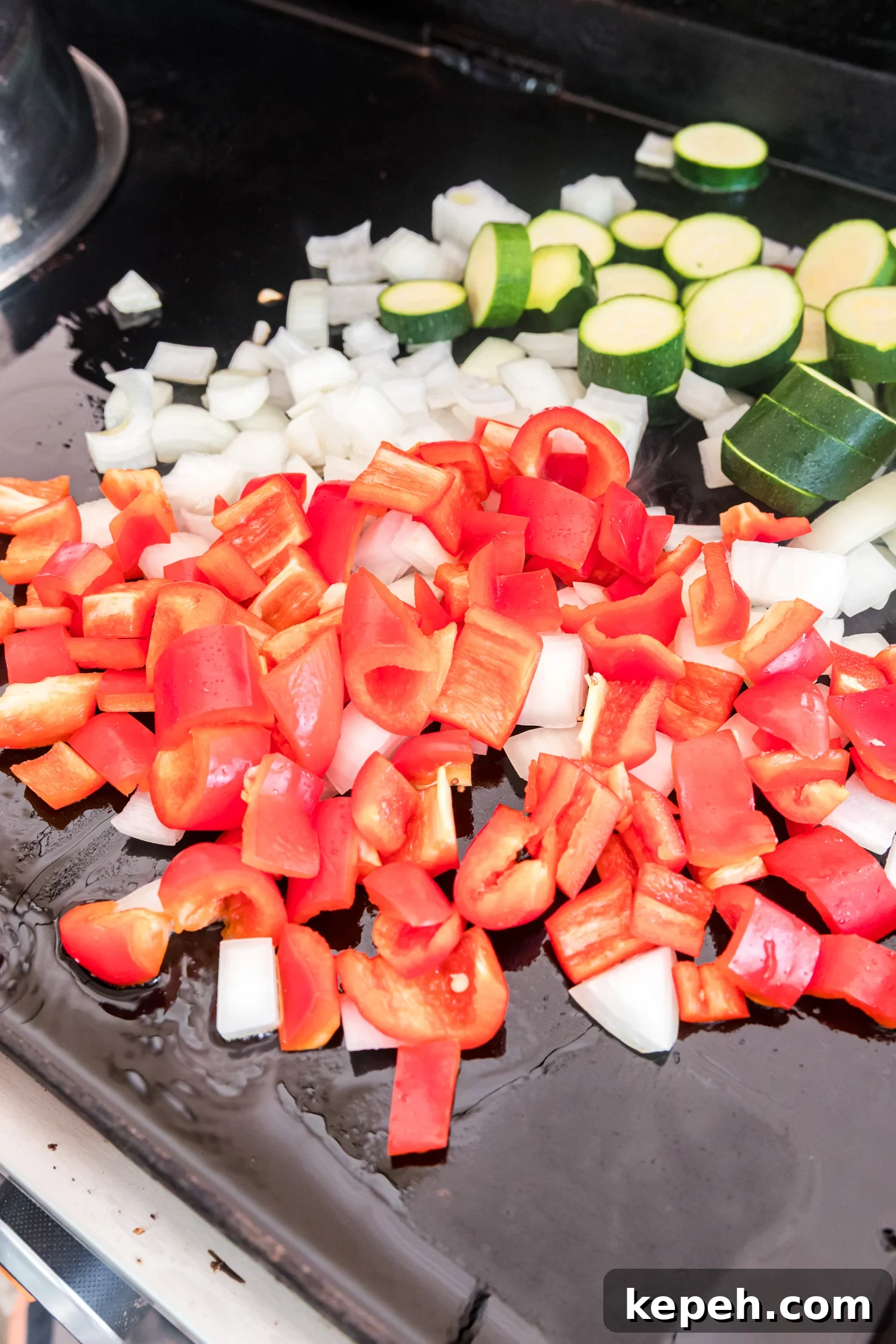 Chopped red bell pepper, diced onion, and sliced zucchini spread across an oiled flat-top griddle.