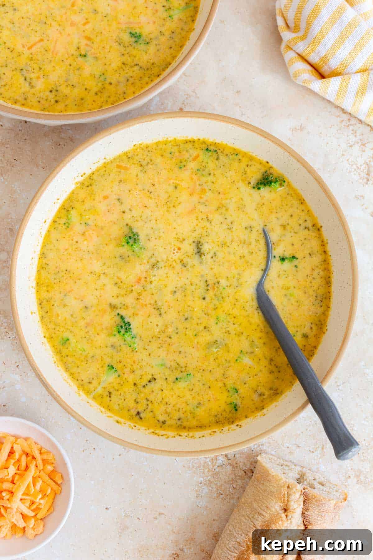 A bowl of Instant Pot broccoli cheddar soup with a grey spoon. Some bread, more cheese, a linen napkin, and another bowl of soup on the side.