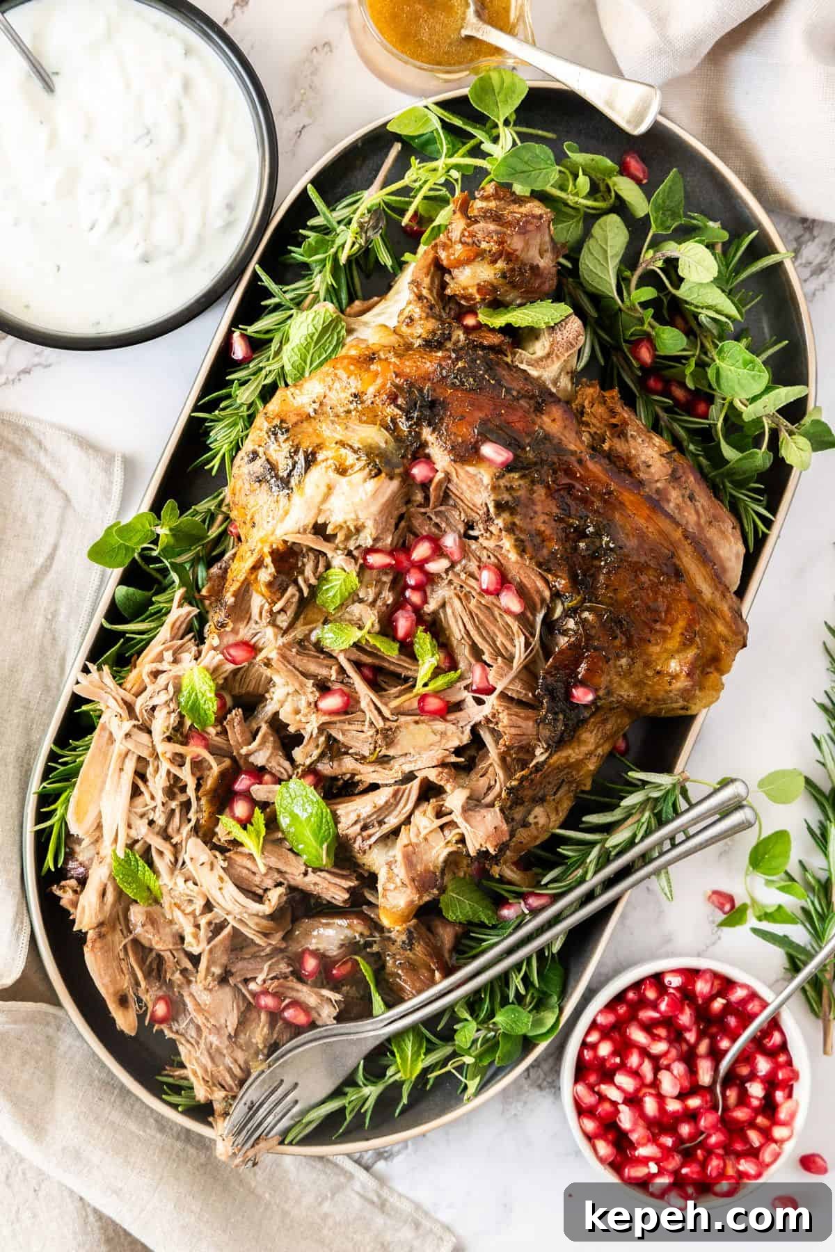 Top Easter Main Courses for a Memorable Holiday Meal 17 Slow Roasted Leg of Lamb, surrounded by fresh herbs and pomegranate, on a grey oval platter, with two forks on the edge.