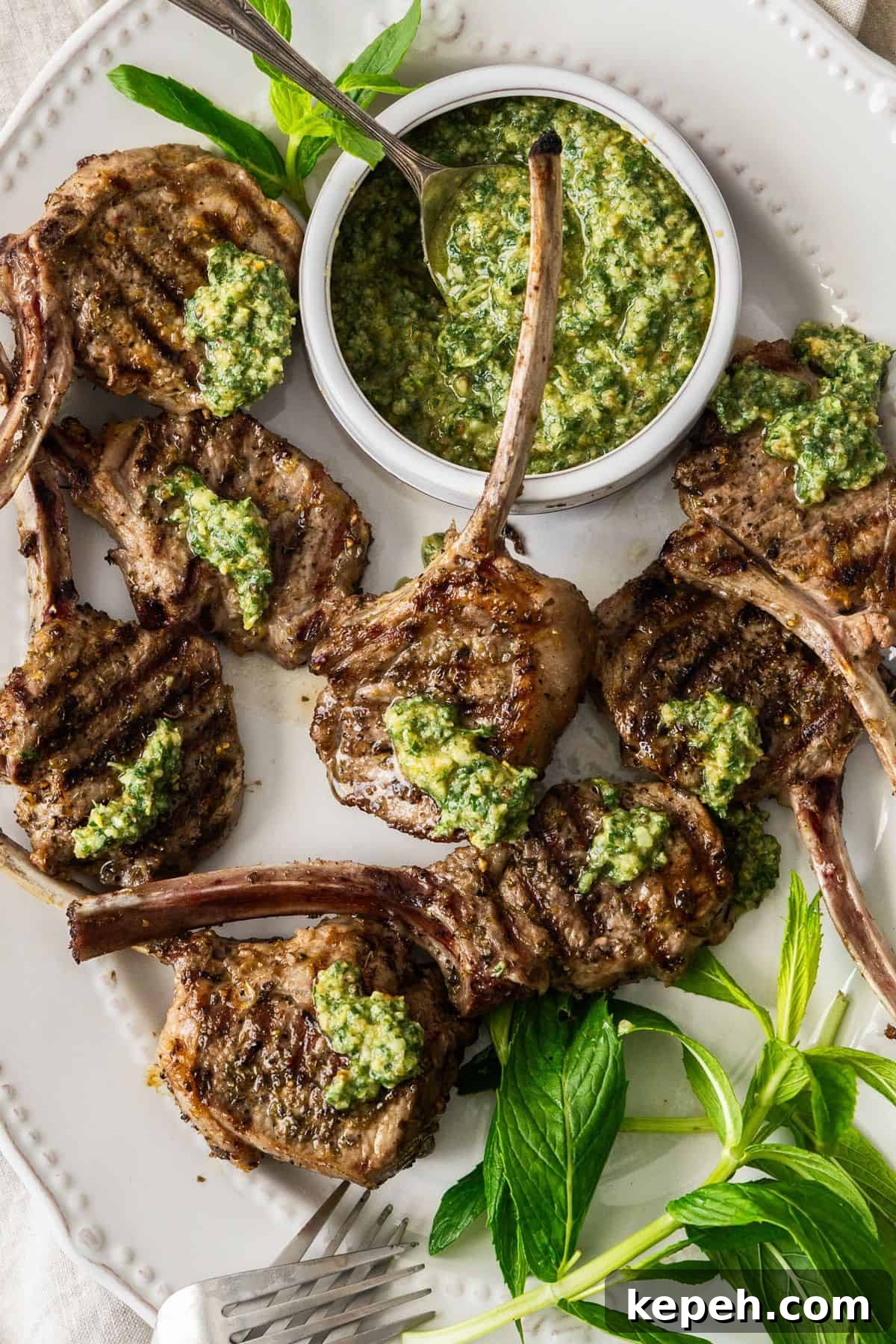 Top Easter Main Courses for a Memorable Holiday Meal 9 Oval white plate with Grilled Lamb Chops, some fresh mint leaves, two forks and a small dish of Mint Pesto.