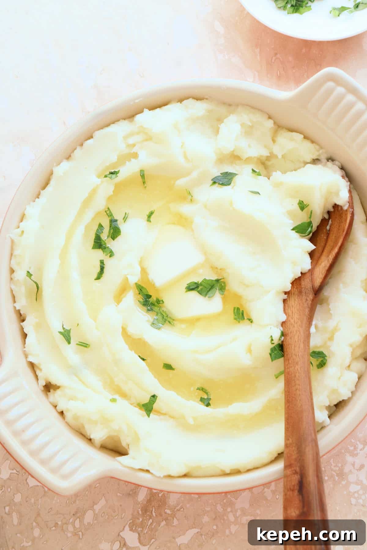 Classic Creamy Mashed Potatoes Recipe for Perfectly Smooth Results 2 Mashed potatoes in a serving dish with a wooden spoon, parsley and melted butter, set on a pink stone board.