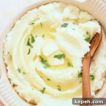 Classic Creamy Mashed Potatoes Recipe for Perfectly Smooth Results 15 Mashed potatoes in a serving dish with a wooden spoon, parsley and melted butter, set on a pink stone board.