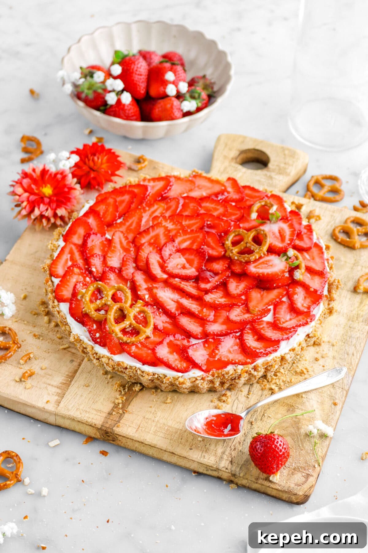 Strawberry Mascarpone Tart with Crunchy Pretzel Crust 20 angled shot of strawberry pretzel tart on wood board wit spoon of jam, strawberries, and flowers around, with bowl of strawberries behind.
