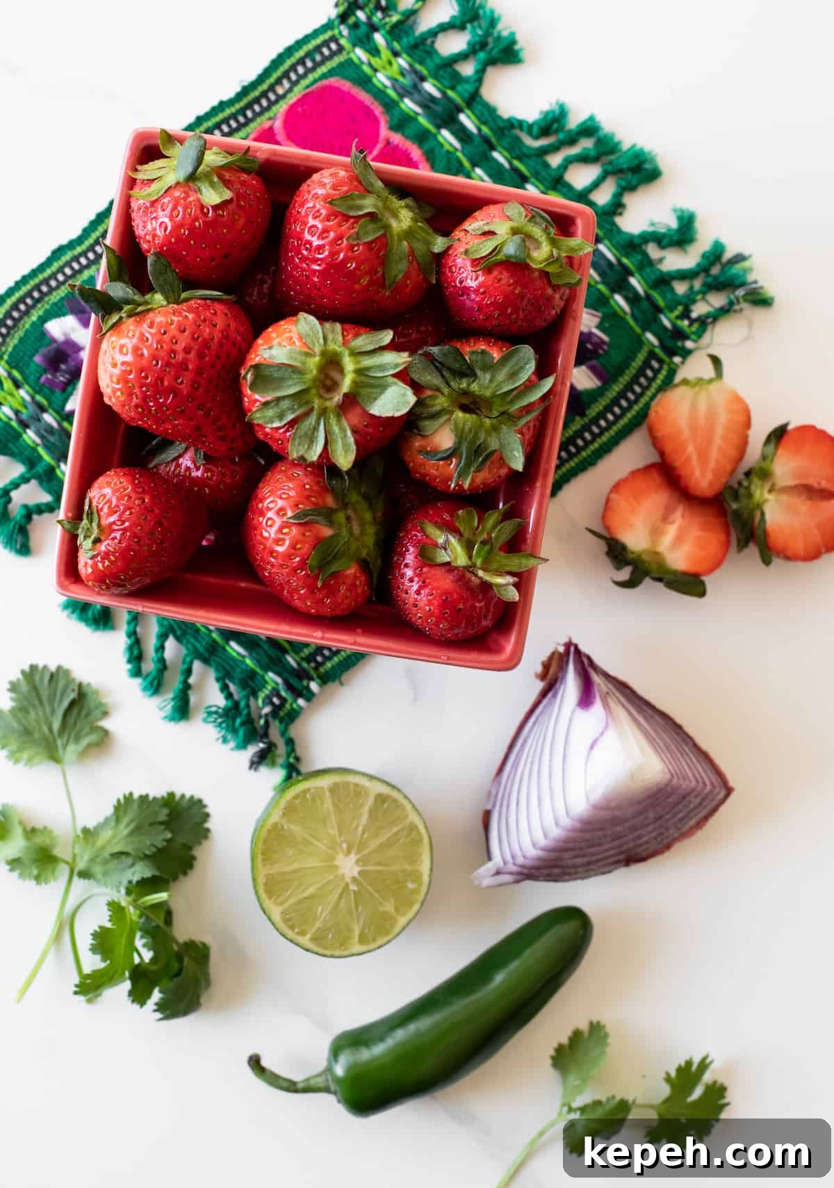 a red container of fresh strawberries, a bunch of fresh cilantro, a quarter of a red onion, a whole fresh jalapeño, and a halved lime on a white table ready to make savory strawberry salsa like a fruit pico de gallo.