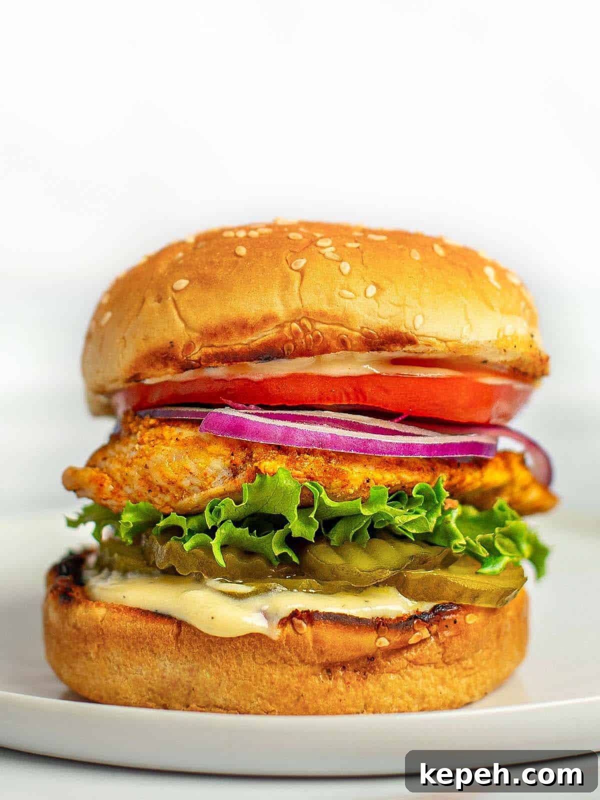 Close up shot of a grilled chicken sandwich stacked with lettuce, tomato, onion, pickles, and honey mustard dressing. 