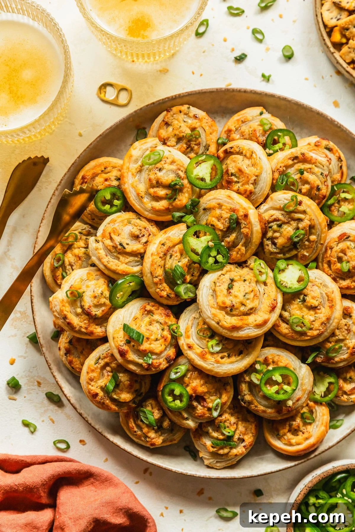 A beige plate of baked jalapeño popper pinwheels on a tan counter next to an orange linen, glasses of beer, and brown bowl of sliced jalapeños.
